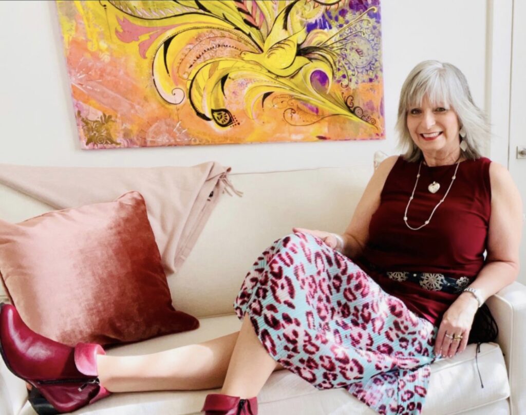 Author Tina is seated on a couch. She has grey hair. There is an abstract painting hanging above couch.